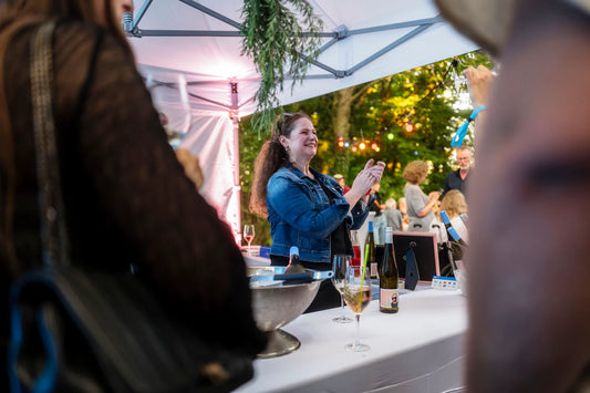 Melie steht in einer blauen Jeansjacke lächelnd unter einem Pavillon auf der Eröffnungsparty, umgeben von Weinflaschen und Gläsern. Im Hintergrund sind bunte Lichterketten und weitere Gäste sichtbar.